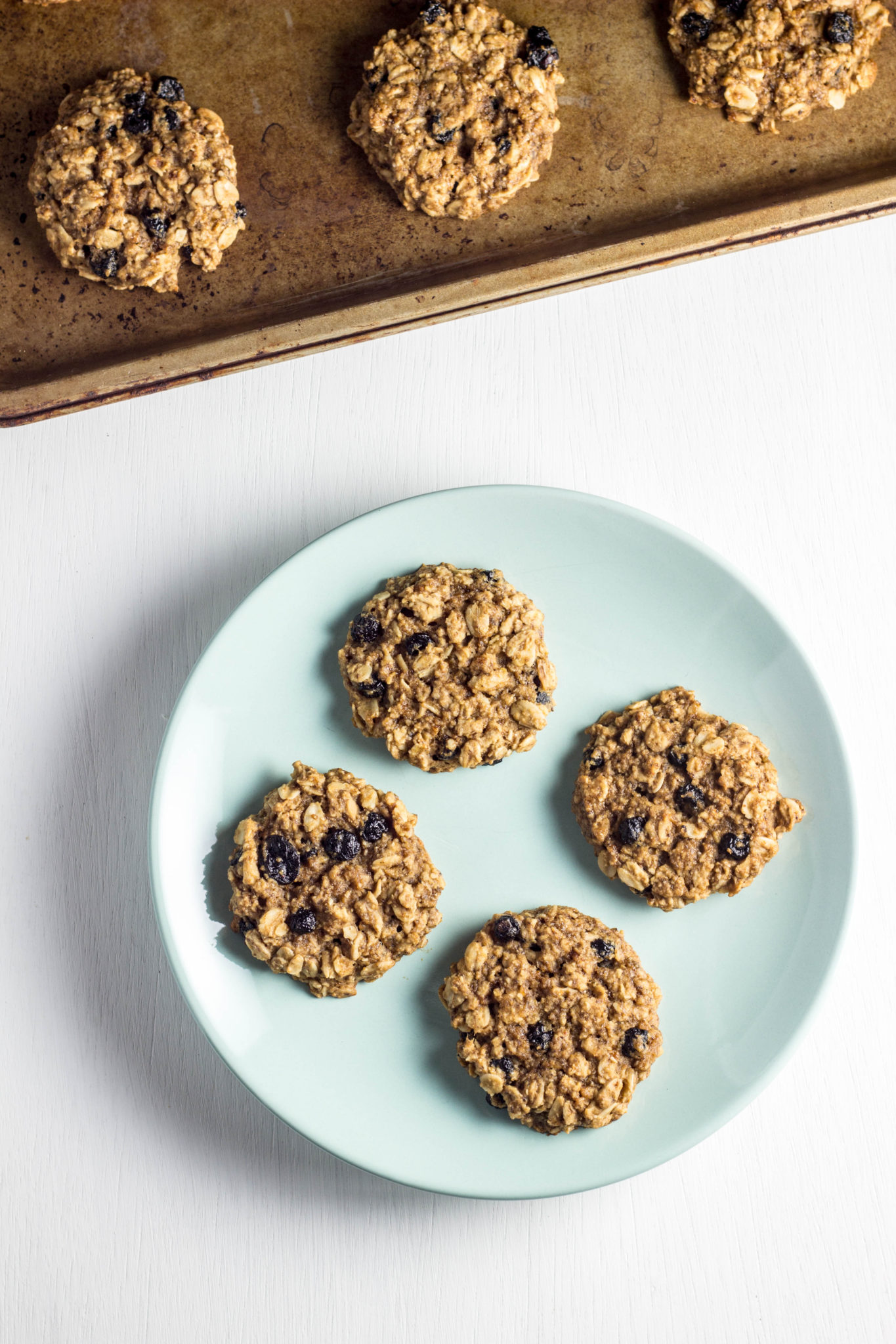 A plate of four Blueberry Oatmeal Cookies against a white background. A baking sheet with more cookies is partially in frame. 