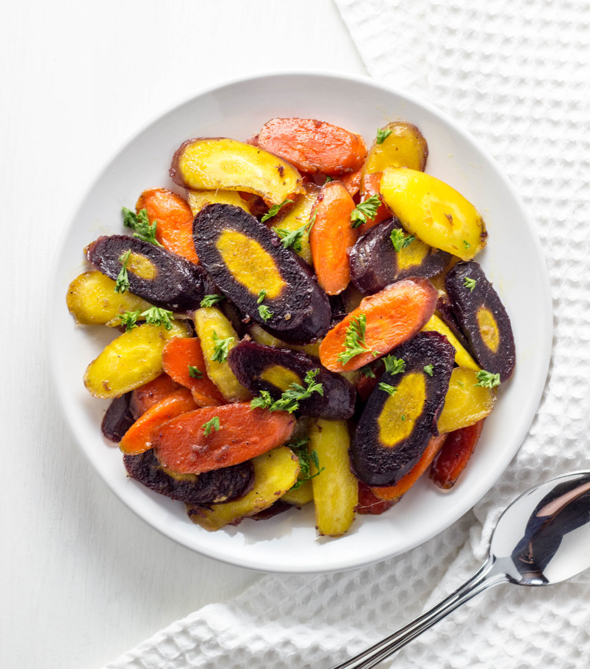 Top view of rainbow carrots on a white plate with a textured towel and spoon to the side. 