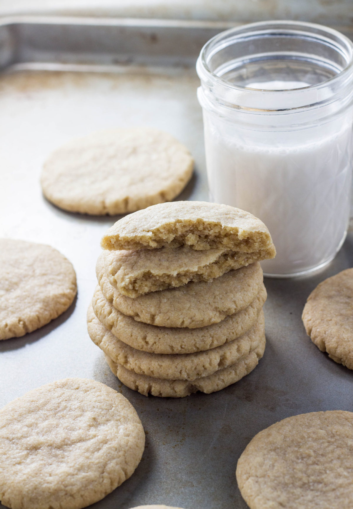 A stack of Easy Vegan Coconut Oil Sugar Cookies with the top cookie broke in half to show interior texture. They're on a baking sheet with other cookies around and a glass of non dairy milk. 