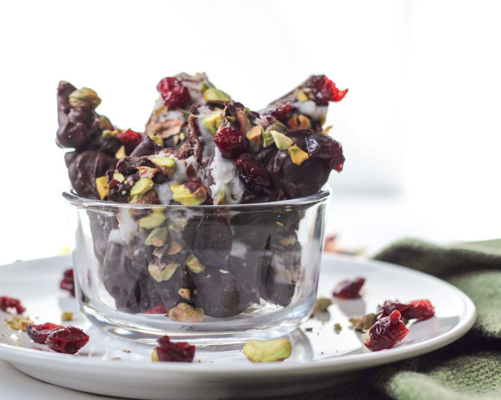 A bowl on a white plate with Dried Cherry Pistachio Chocolate Bark in it. There are pistachios and cherries scattered on the plate around the bowl. 