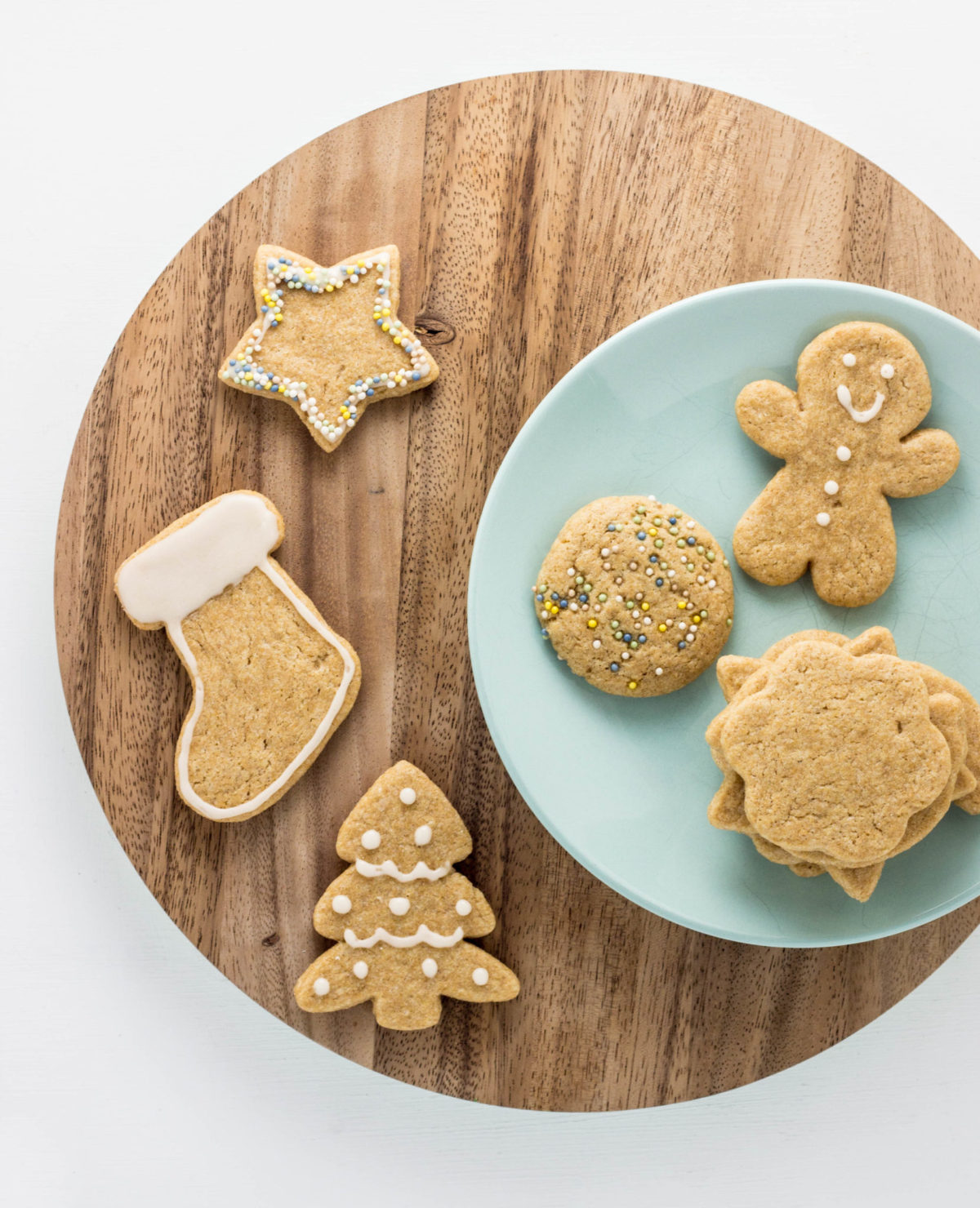 Top view of cut out sugar cookies. Some are on an aqua colored plate and sitting on a round wooden cutting  board while some are on the board to the side of the plate. 
