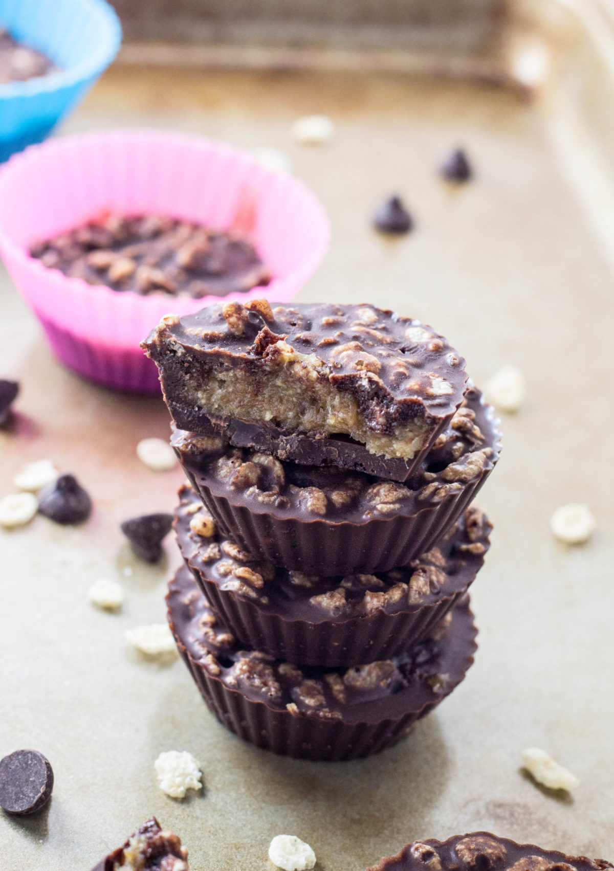 Side view of a stack of four Vegan 100 Grand Candy Bar Cups with the top cut in half to show filling. There are rice cereal and chocolate chips around the stack. 