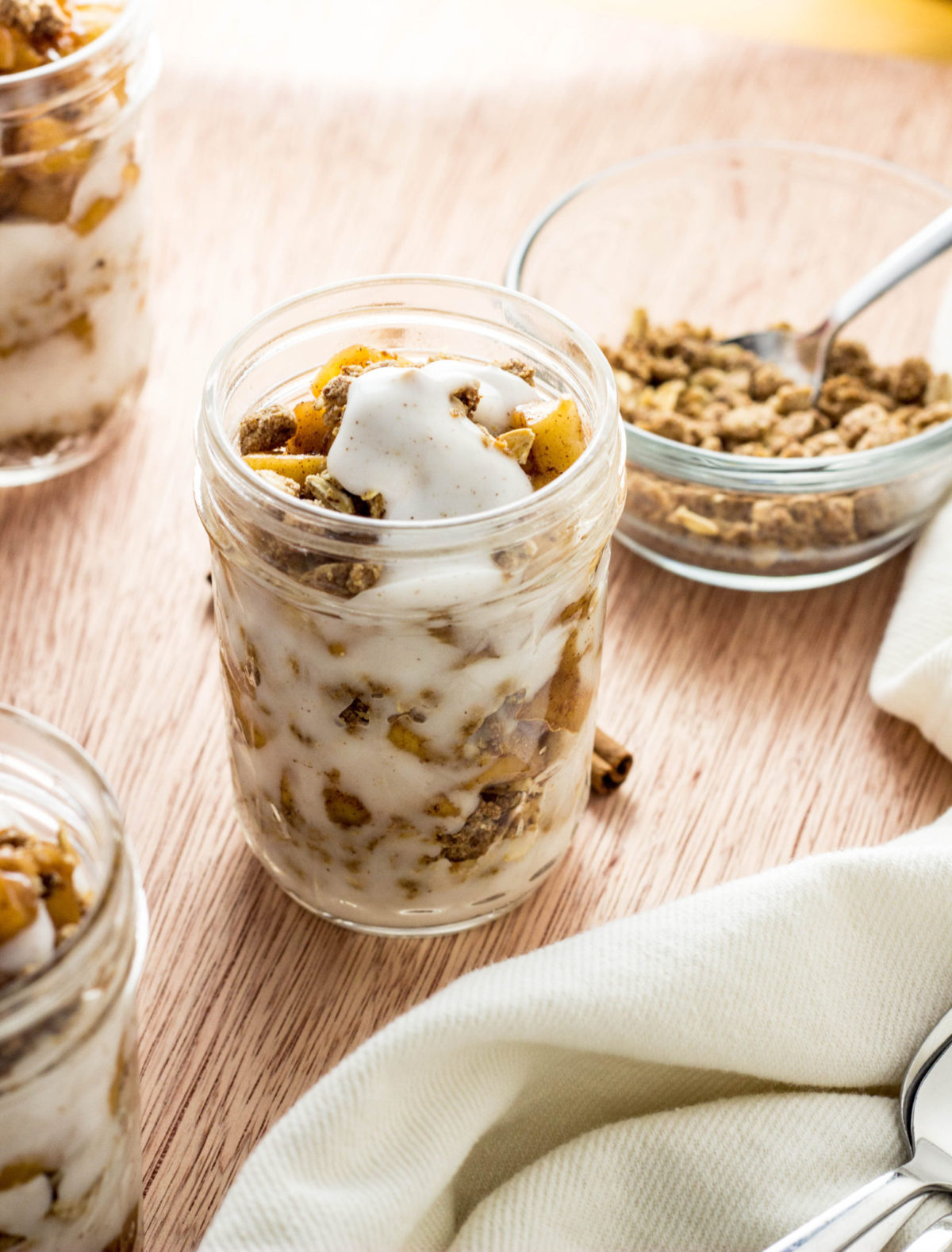 A 45 degree angle top view of Vegan Sautéed Apple Yogurt Parfait in a clear jar. There are other jars partially visible and a bowl of granola n the background 