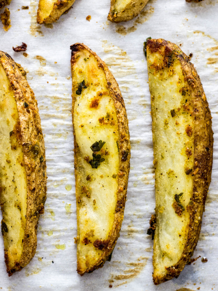 Close up top view of Vegan Cheesy Herbed Potato wedges on white parchment paper.