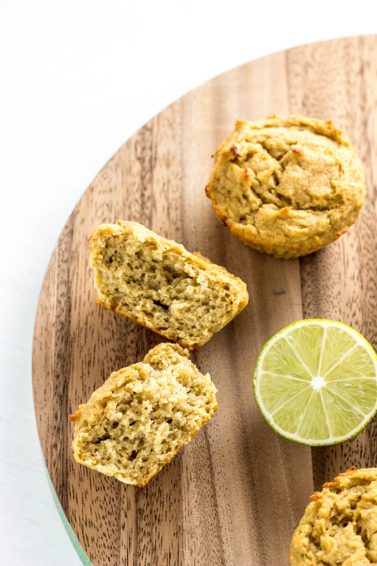 Vegan Lime Avocado Muffins on a round wooden cutting board with a wedge of lime. One the the muffins is cut in half turn on the side to show the interior texture. 