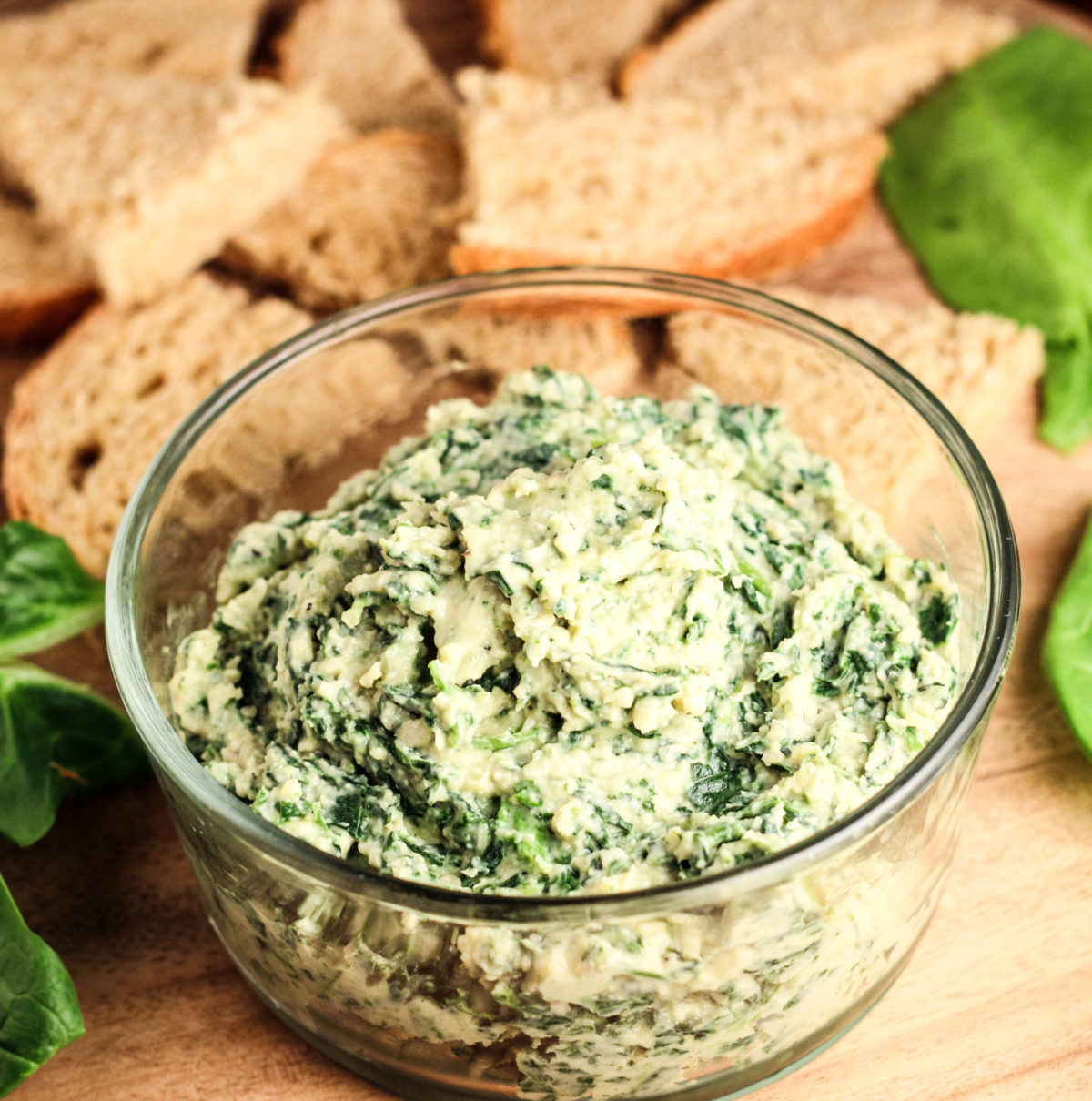 Close up 45 degree view of bowl of Vegan Spinach White Bean Dip. In the background there are pieces of bread and spinach leaves. 