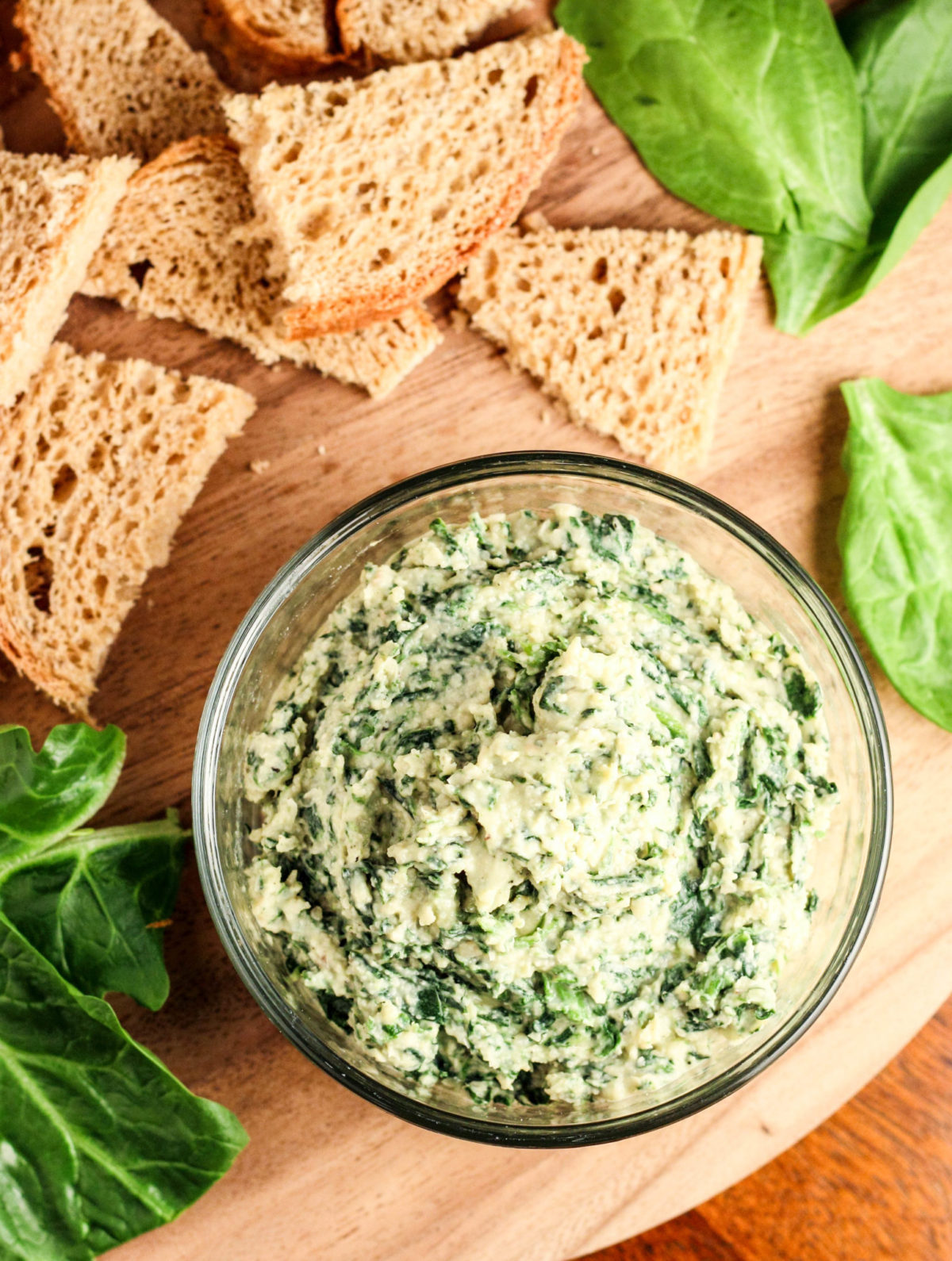 Top view of a bowl of Vegan Spinach White Bean Dip with pieces of bread and spinach around. 