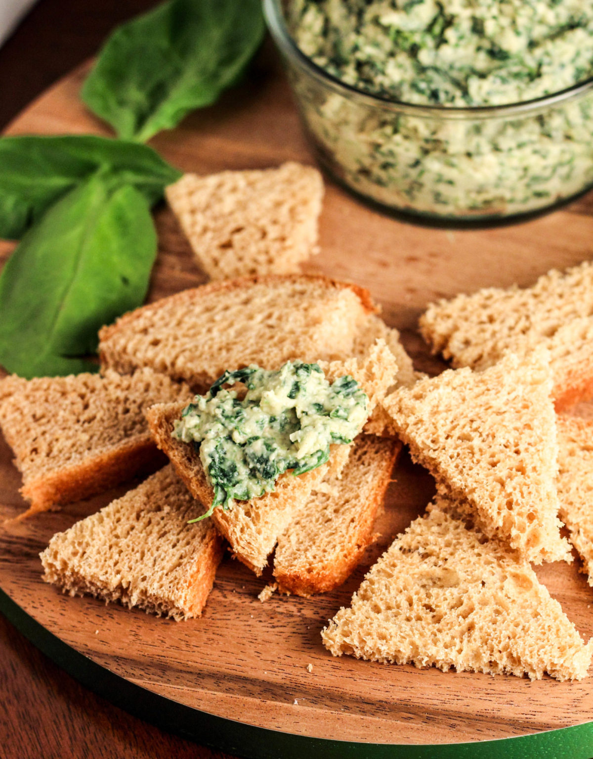 Dip spread on a toast point among other pieces of toast on a wooden cutting board. There is a bowl of dip and spinach leaves in the background. 