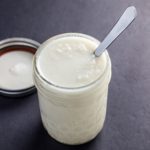 A small jar of coconut butter with a spoon sticking out of the jar on a black background.