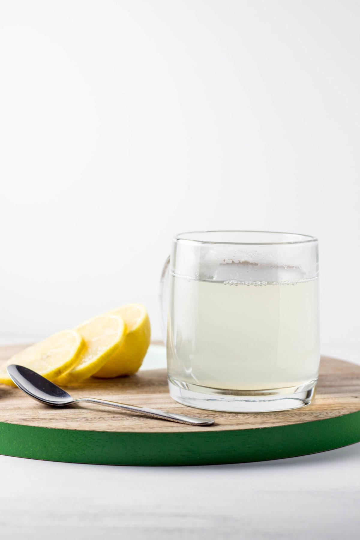 Side view of Ginger Lemon Tea in a clear mug on a wooden cutting board. There is a spoon and lemon slices to the side. 