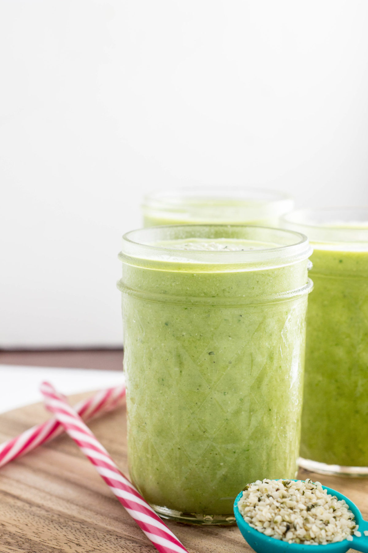 Side view of of a glass of Kid Friendly Green Smoothie  with straws and a spoon of hemp seeds. 