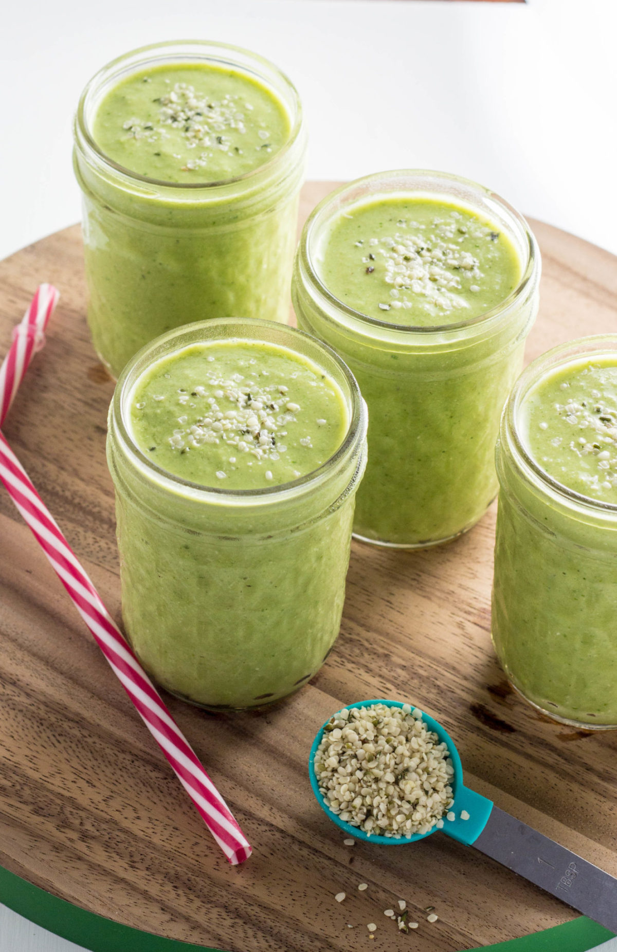 Four glasses of Kid Friendly Green Smoothie at a 45 degree angle on a round wooden cutting board. There is also straws and a spoon filled with hemp seeds. 