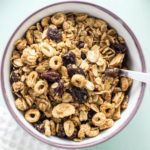 Top view close up of Oatmeal Raisin Cookie Granola in a white bowl with purple rim and a utensil handle visible on the right side.
