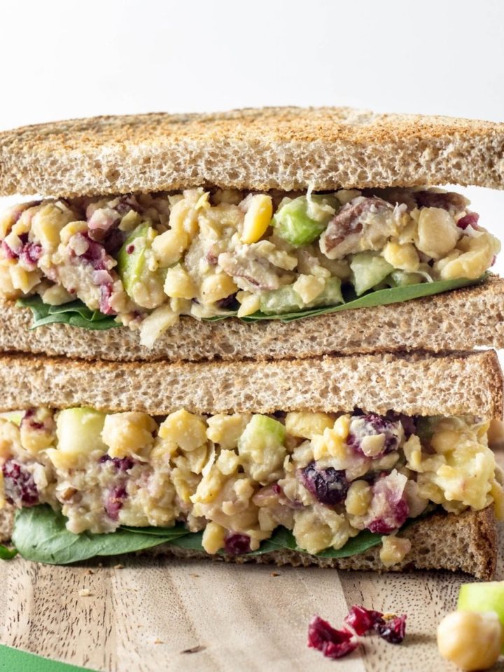 Close up side view of two Vegan Cranberry Apple Chickpea Salad sandwich halves stacked on a wooden board.