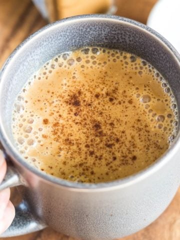 Vegan Warm Spiced Pumpkin Milk in a gray mug with a hand on the handle. The background is wood and a small white bowl with spices is beside the mug.