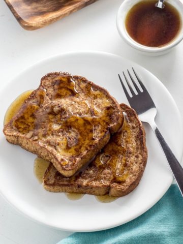 Two slices of Vegan Gingerbread French Toast on a white plate with a fork and a light blue cloth napkin beside the plate. There is a white dipping bowl with cardamom syrup to the side of the plate.