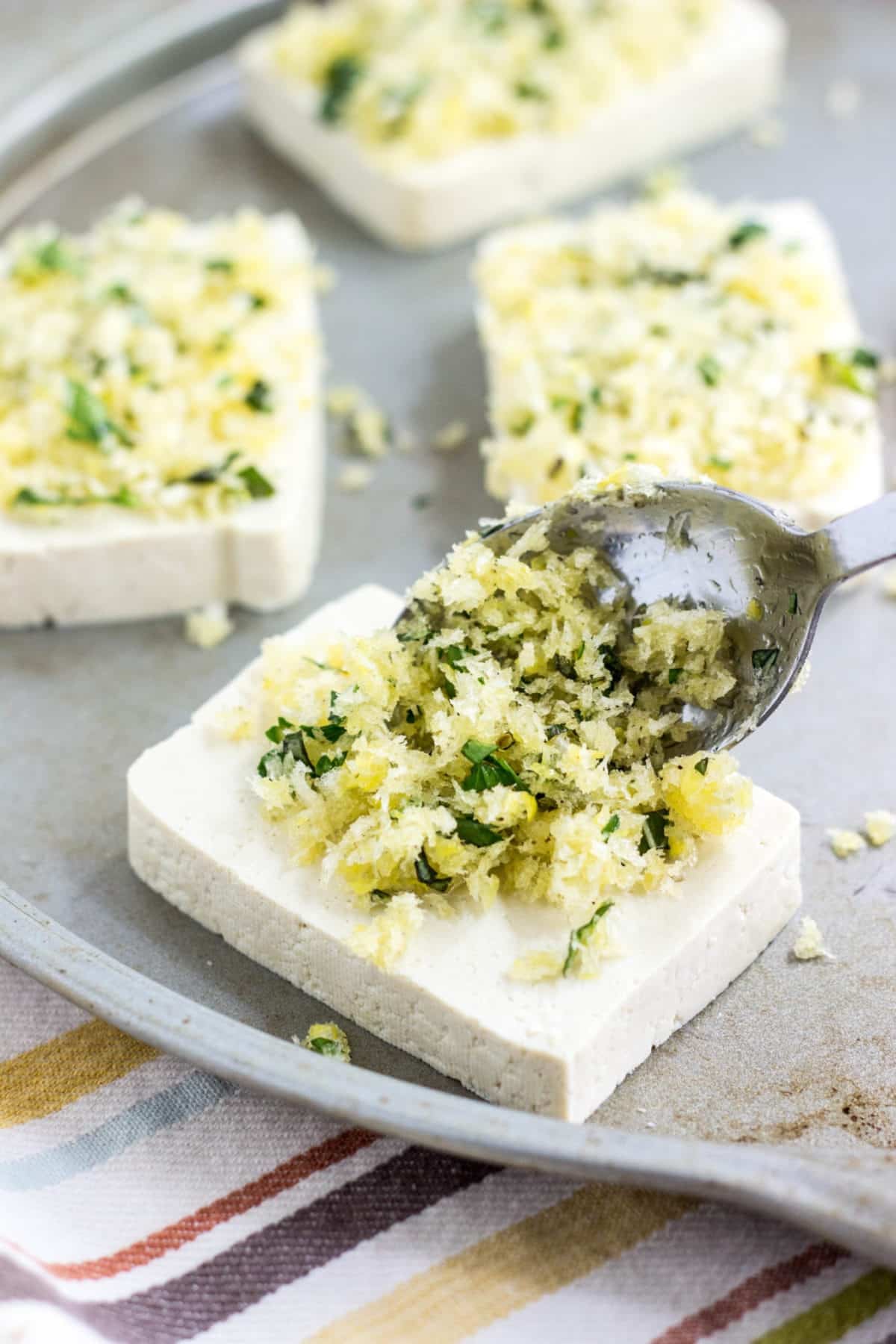Tofu on a baking sheet with a spoon place bread crumb and herb mixture on top.