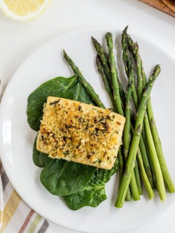 Top view of Baked Lemon & Herb Crusted Tofu on a bed of spinach and a side of asparagus on a white plate