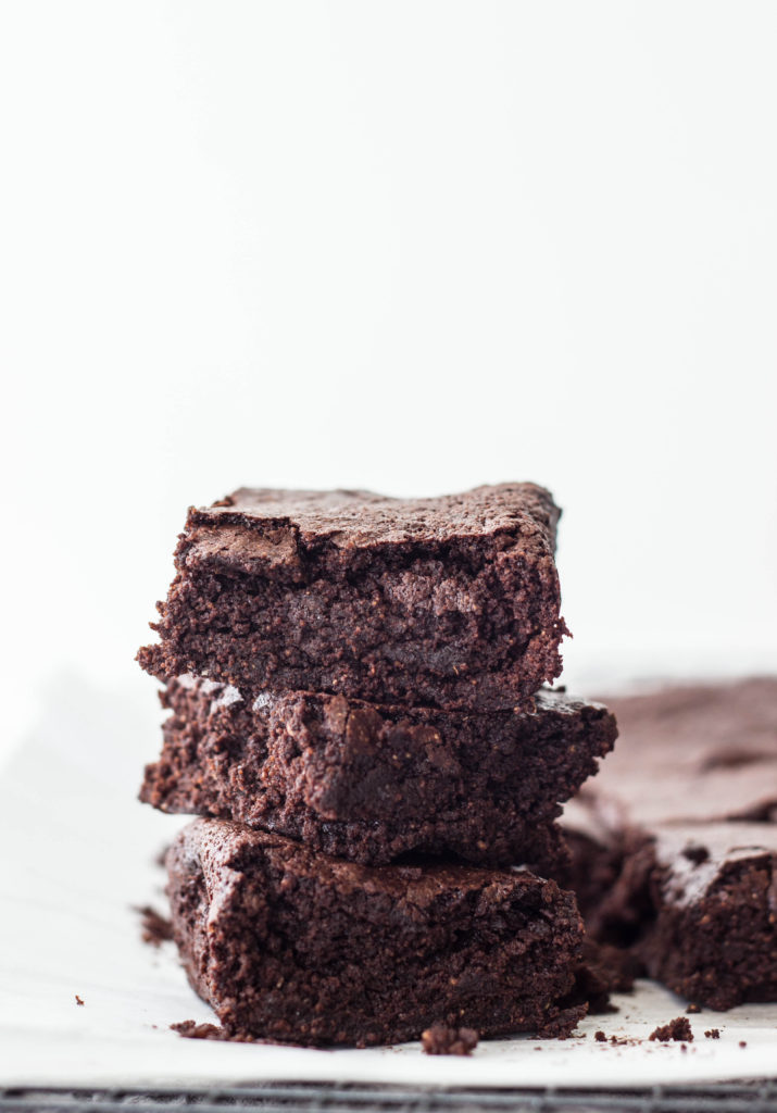 Close up side view of a stack of three Vegan Rye Crinkle Brownies on white parchment paper with other brownies in the background. 