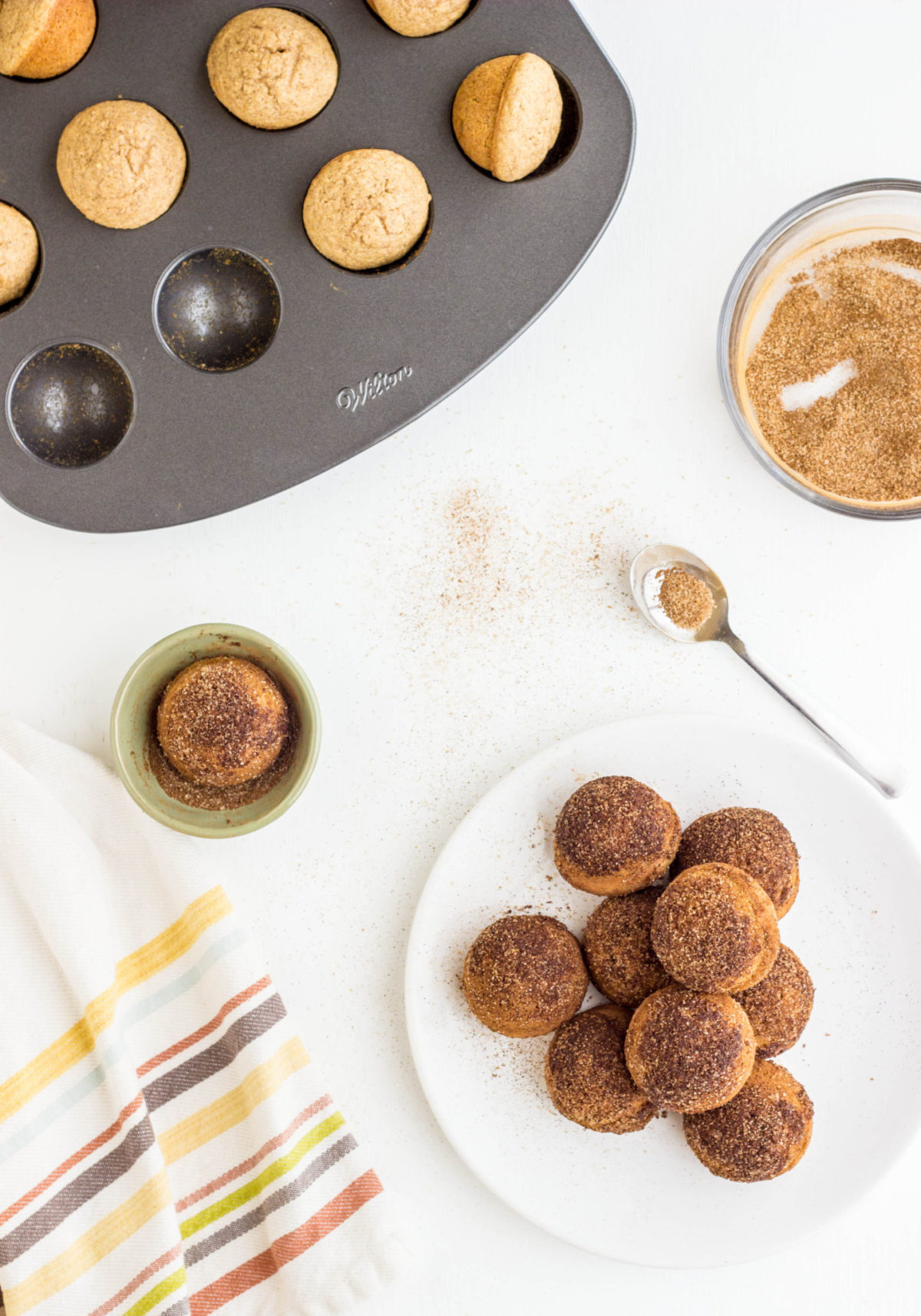 Top view of Doughnut Holes on white plate with bowls and pan around. 