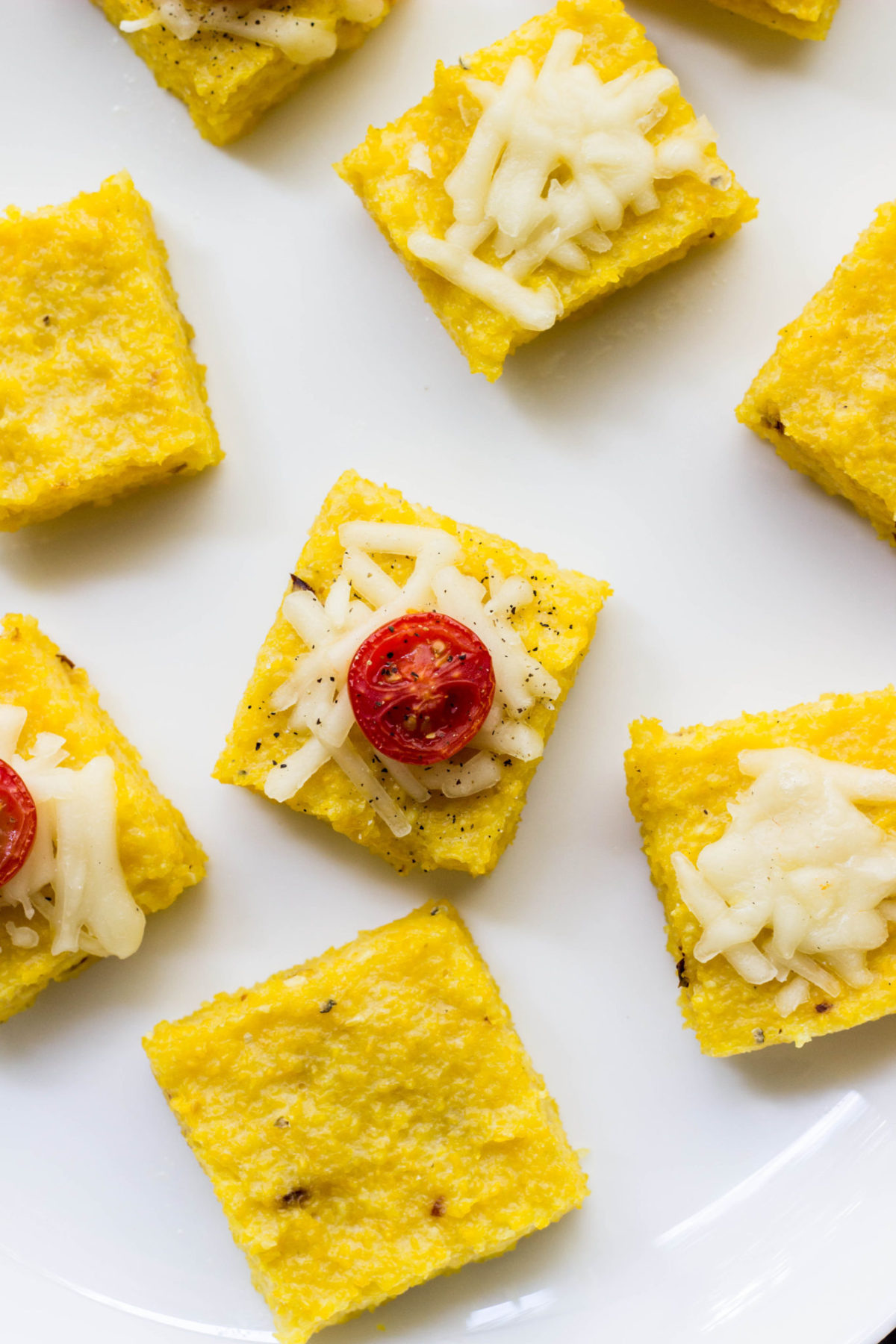 Vegan Cheesy Polenta Bites on a white plate. Some are topped with vegan cheese and tomato. 