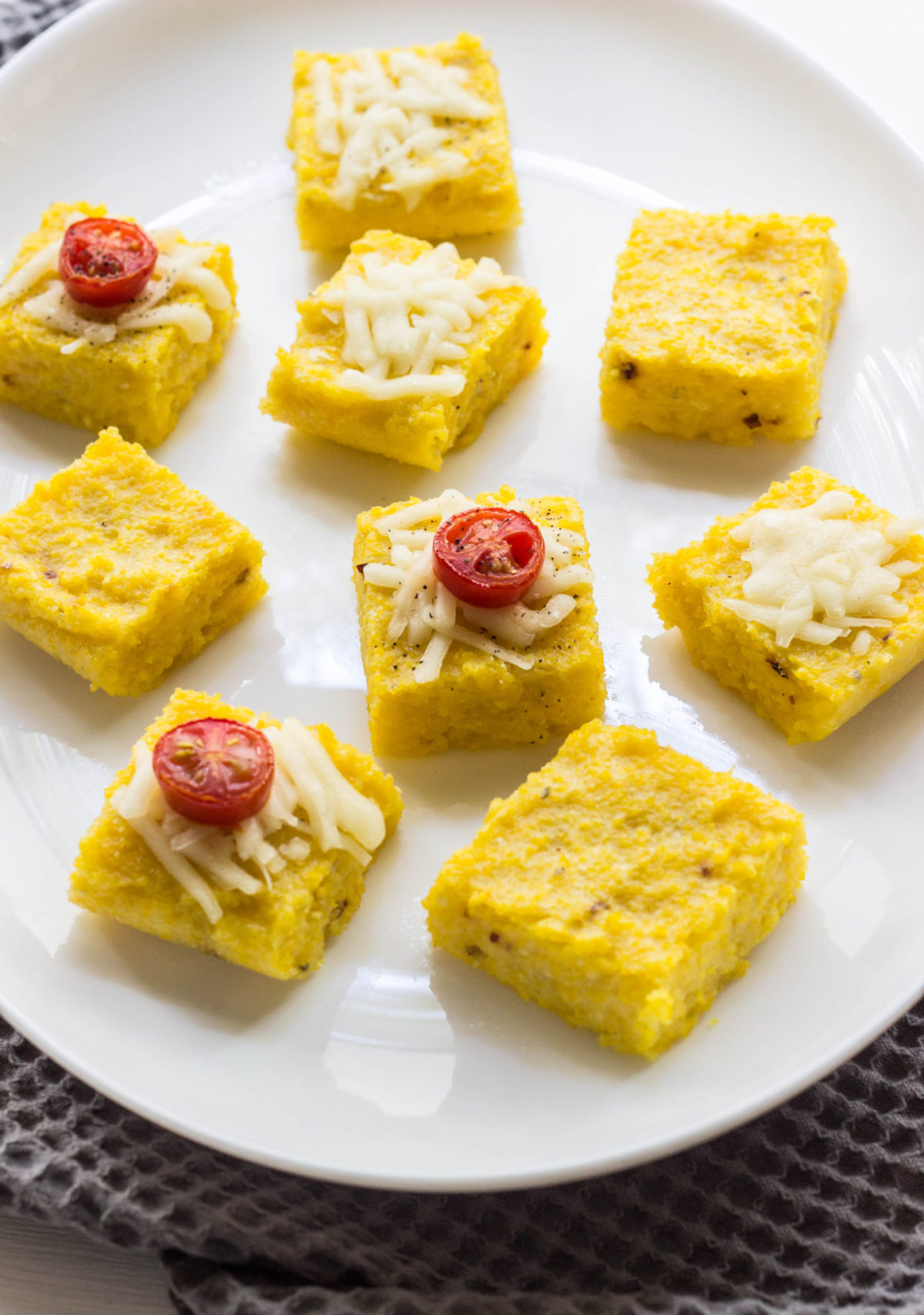 Polenta bites on a white plate with vegan cheese and tomato toppings. 