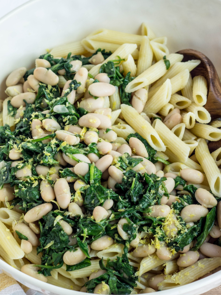 Close up of White Beans and Greens Pasta in cream bowl with wooden utensils.