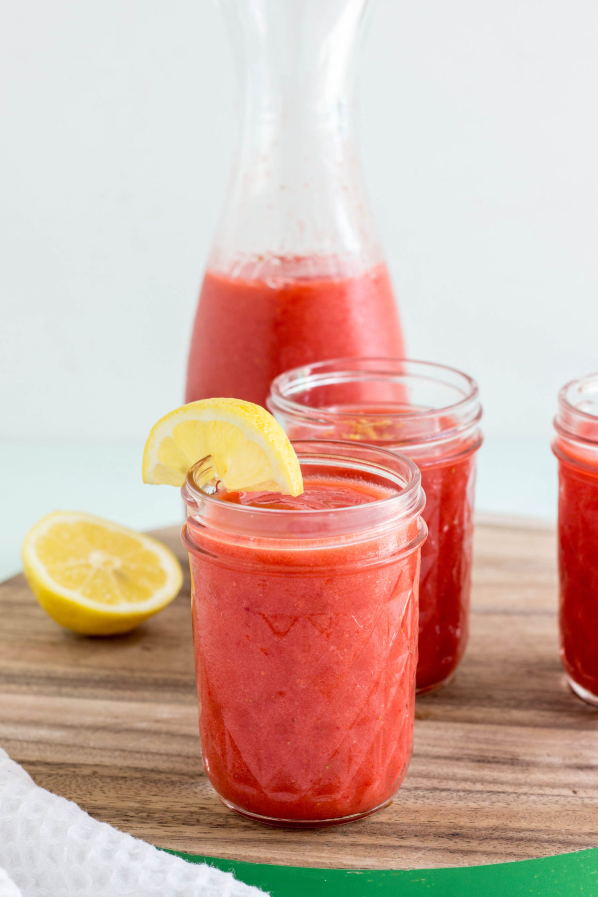 Side view of three glasses of lemonade on a wooden board. In the background is a carafe of lemonade and a wedge of lemon. 