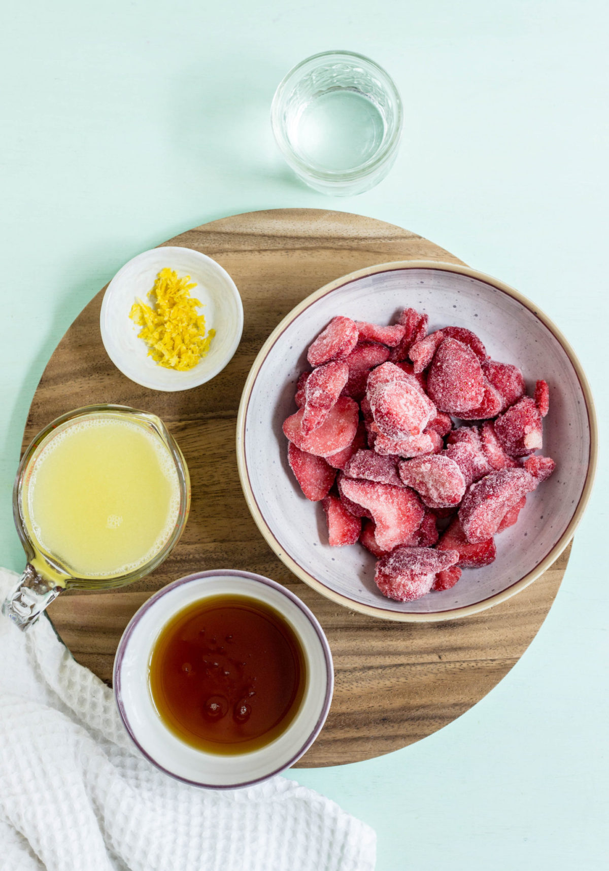 Top view of bowls of ingredients used to make strawberry lemonade including frozen strawberries, lemon juice, zest, maple syrup and water. 