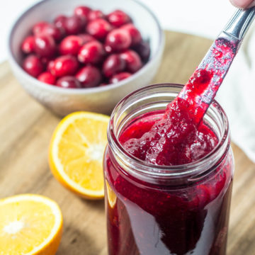 Side view of a jar of Simple Cranberry Orange Jam with a knife lifting jam out of it. In the background are two orange halves and a bowl of fresh cranberries.