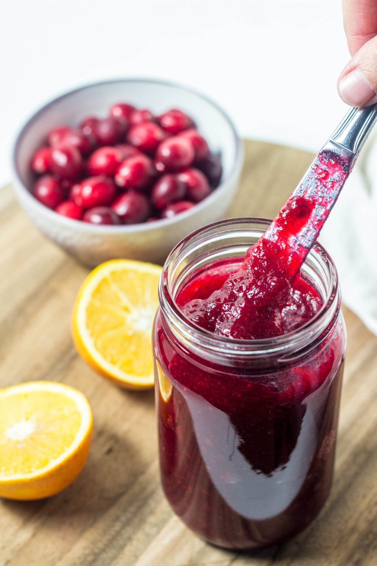 Side view of a jar of Simple Cranberry Orange Jam with a knife lifting jam. In the background are two orange halves and a bowl of fresh cranberries.