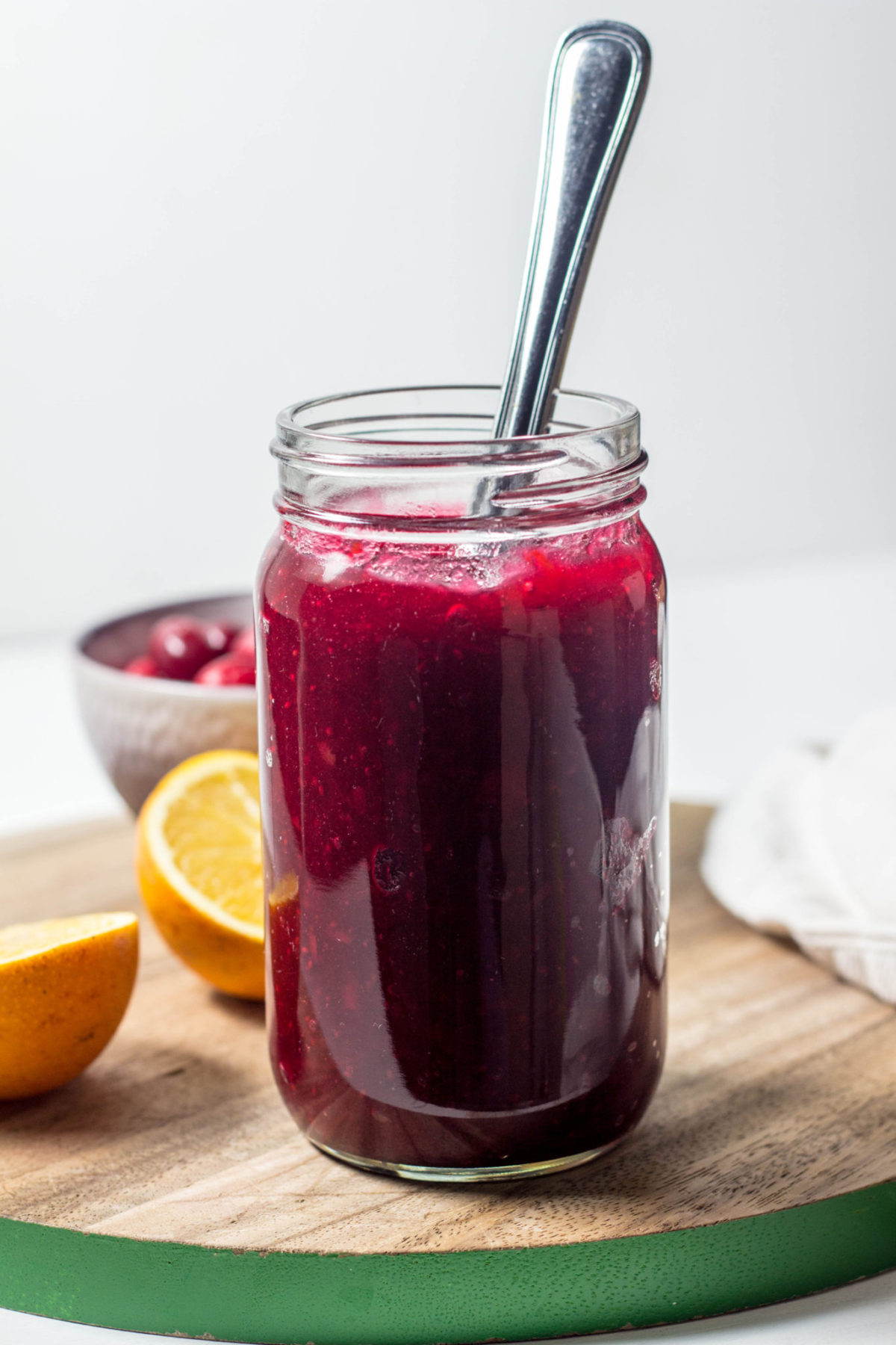 Side view of a jar of Simple Cranberry Orange Jam with a knife sticking out of it. In the background are two orange halves and a bowl of fresh cranberries.