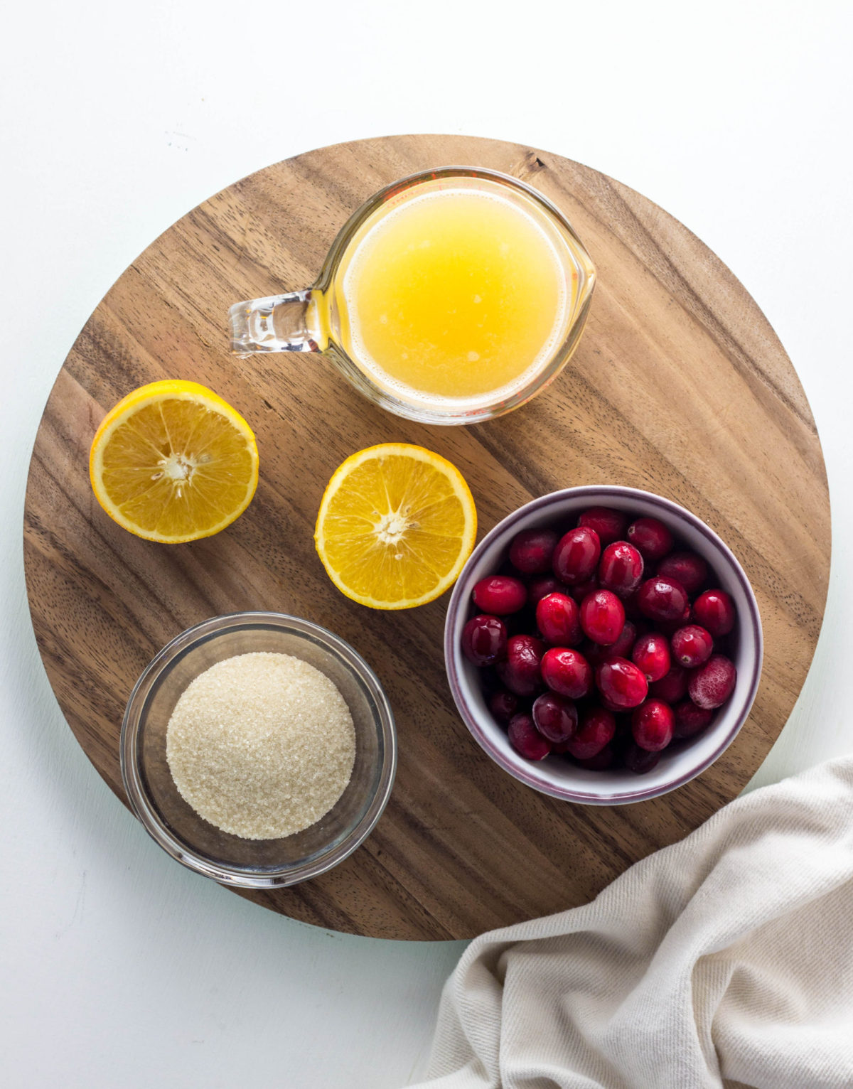 Top view of ingredients needed to make cranberry orange jam - a bowl of cranberries, orange halves, orange juice and sugar on a round wooden board. 