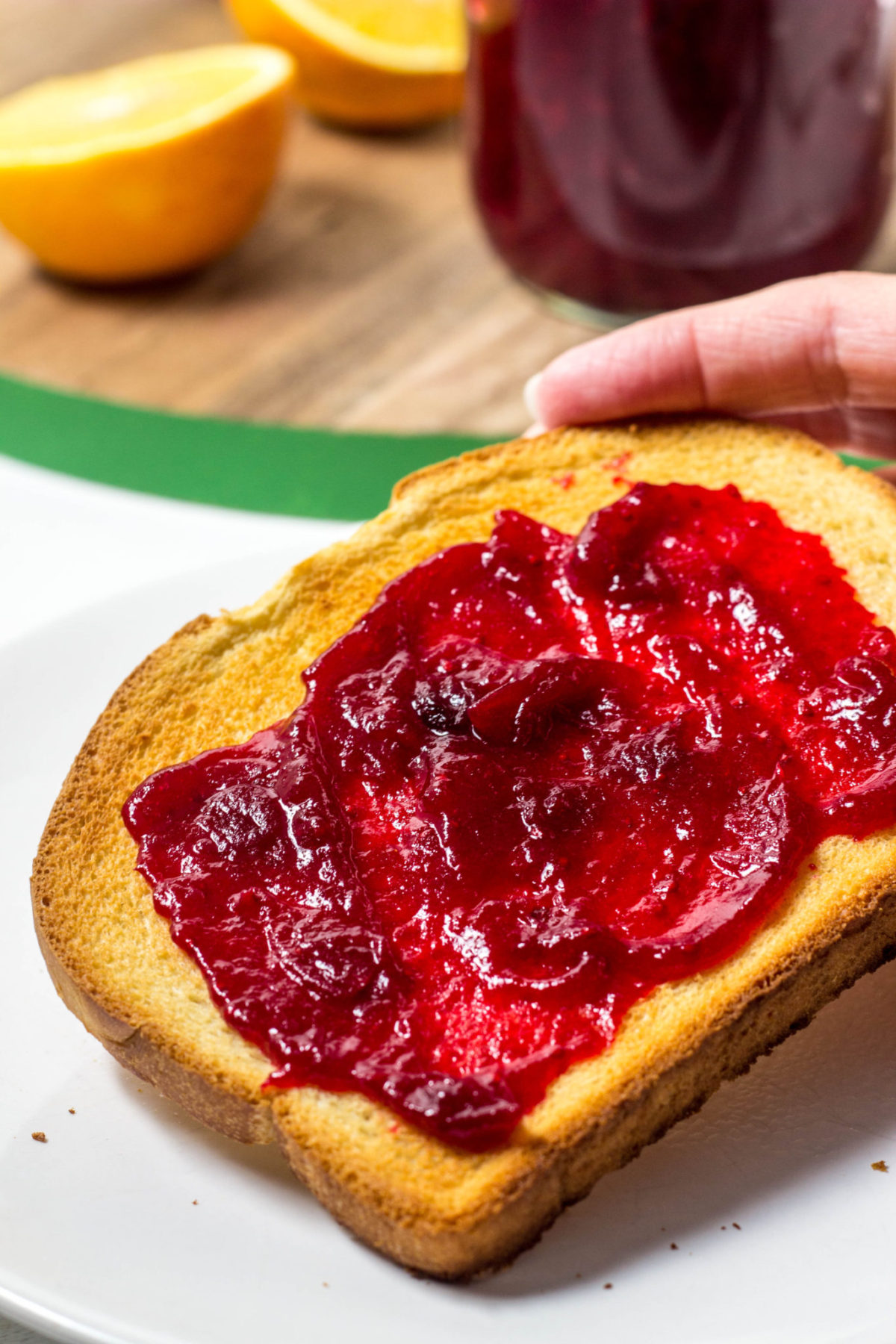 There is a slice of toast with cranberry jam slathered on it being held by a hand. In the background there is a cutting board with orange halves and a jar of jam. 