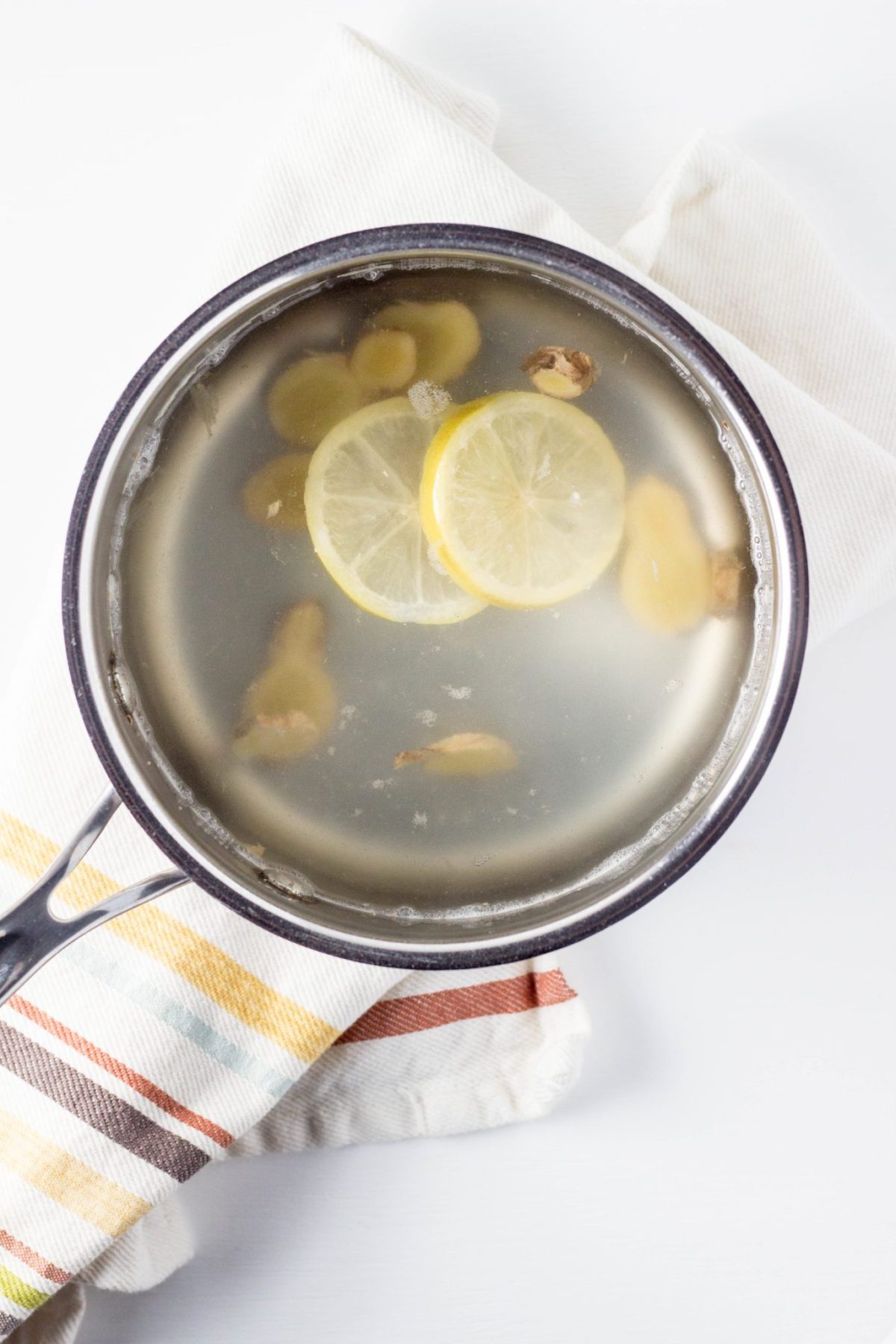 Top view of tea in a sauce pan with lemon slices and slices of ginger with a striped towel. 