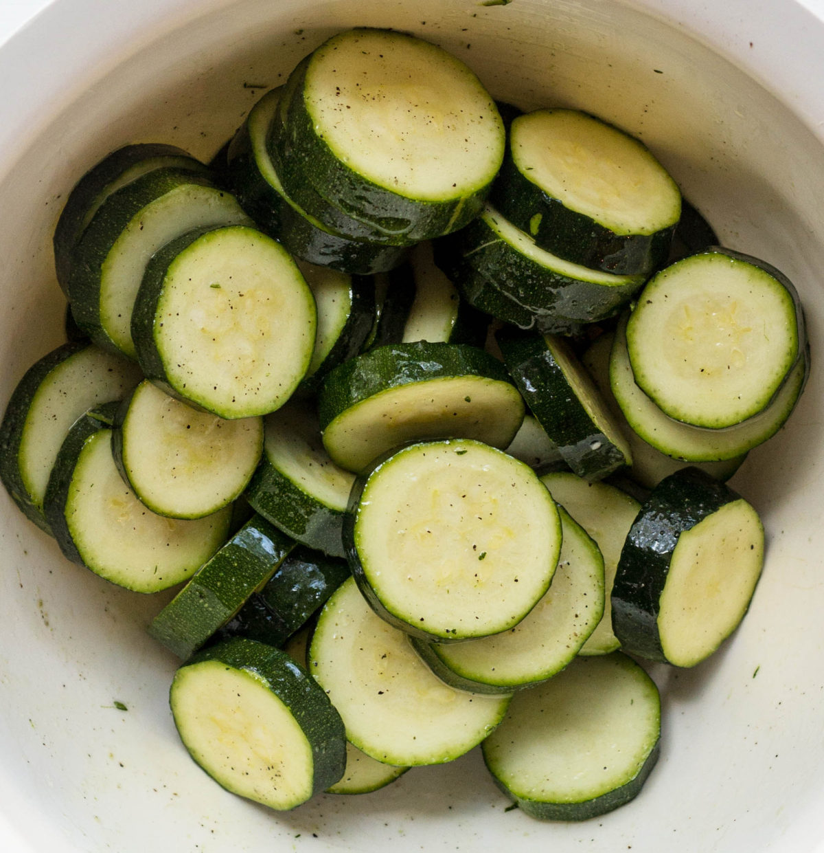 Close up of raw zucchini slices coated in olive oil and seasonings in a white bowl. 