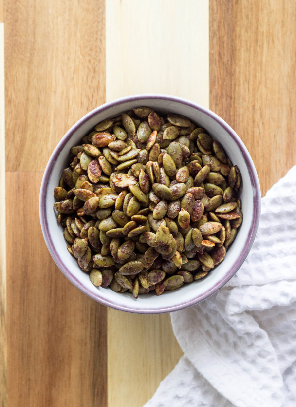 Sweet and Salty Roasted Pumpkin Seeds in a small white bowl with purple rim against a wood background. 