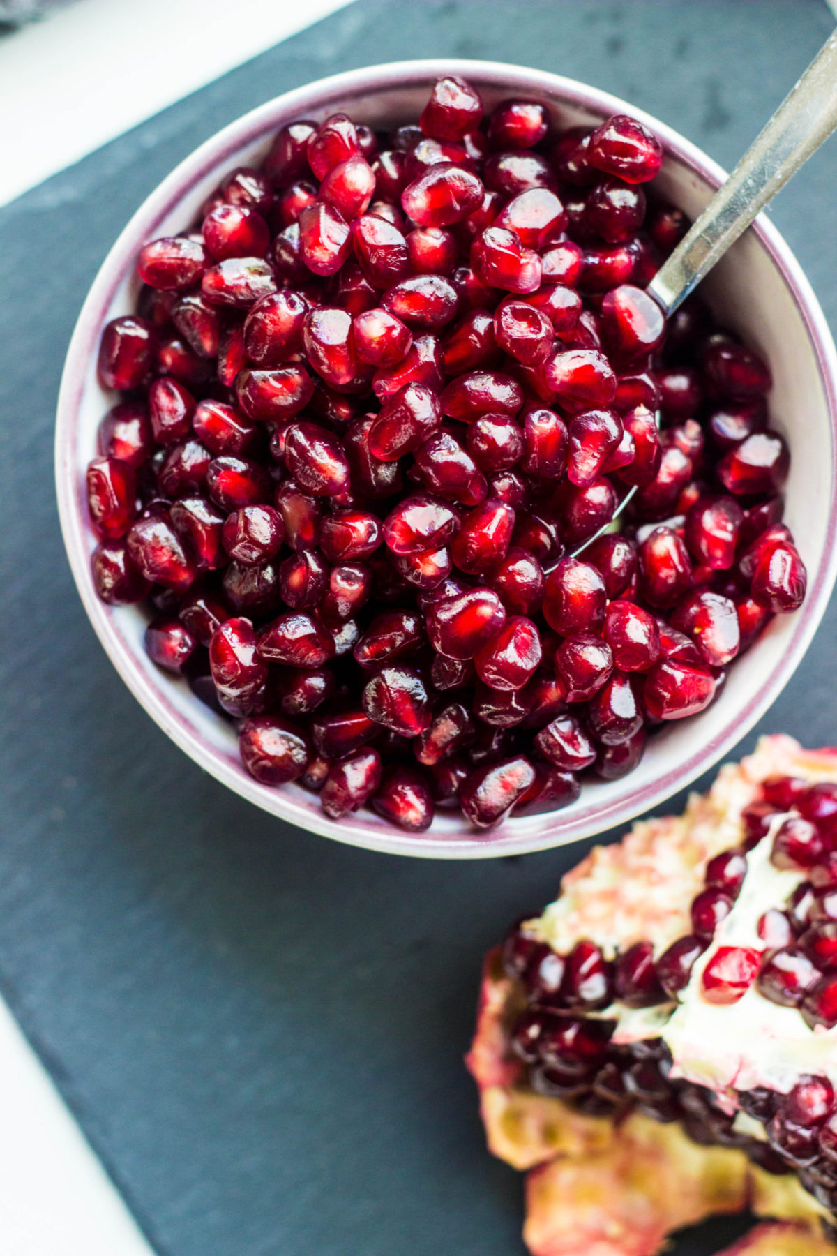 Close up of pomegranate arils in a white bowl with purple rim with a spoon. 