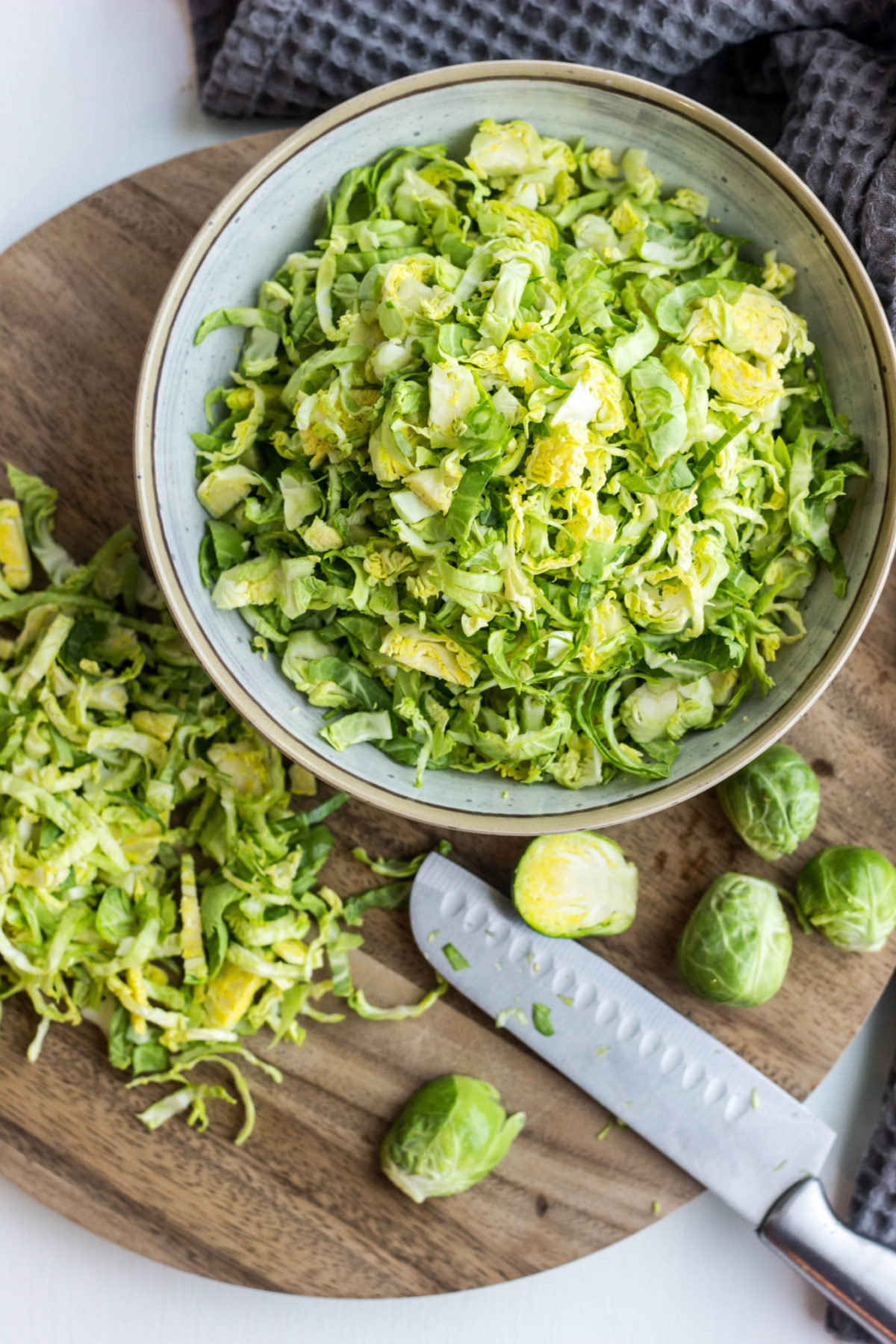 Thinly sliced Brussels sprouts in bowl on a wooden cutting board with knife and more Brussels sprouts.