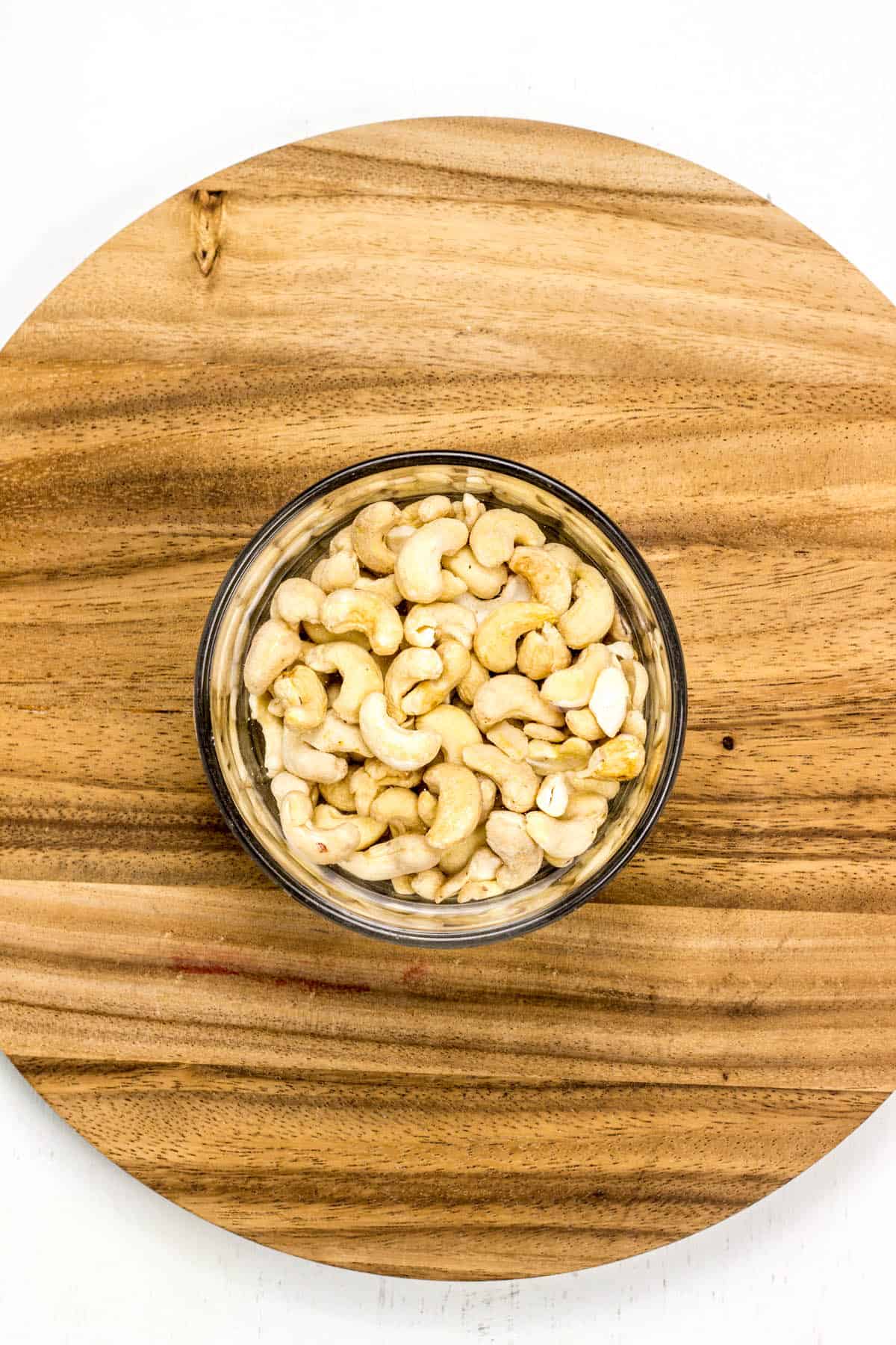Top view of raw cashews in a small bowl on a wooden board.
