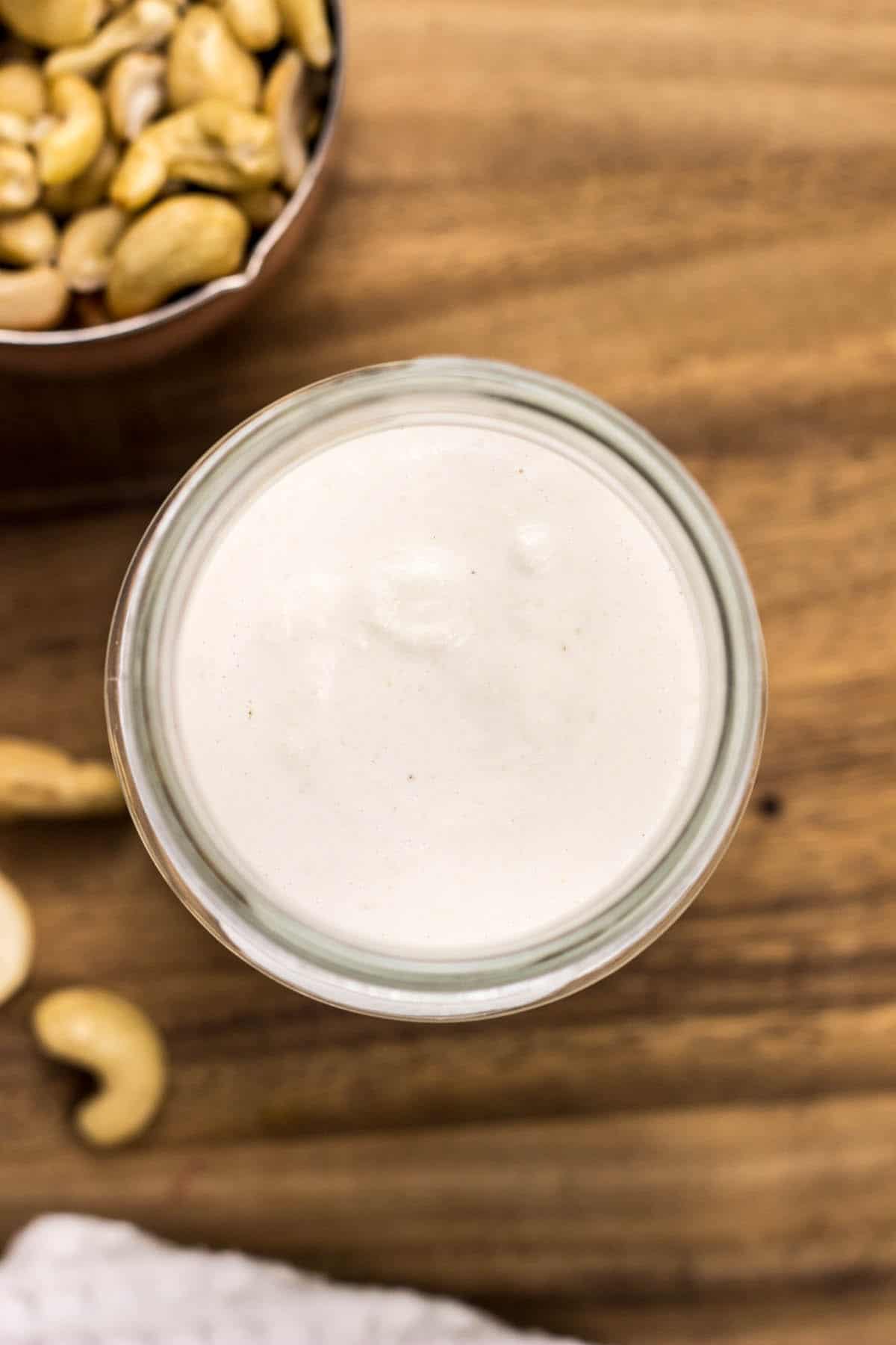 Top view of a small jar of Cashew Cream on a wooden board with a few cashews surrounding.