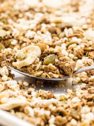 Close up of a spoon of granola resting on a baking sheet of more granola.
