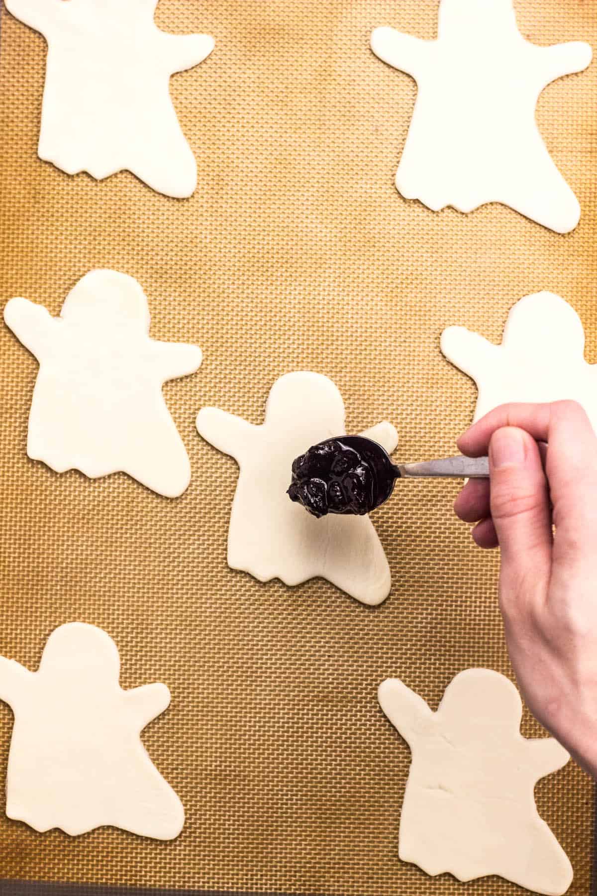 Top view of puff pastry cut into ghost shapes with a spoon placing blueberry filling on one.