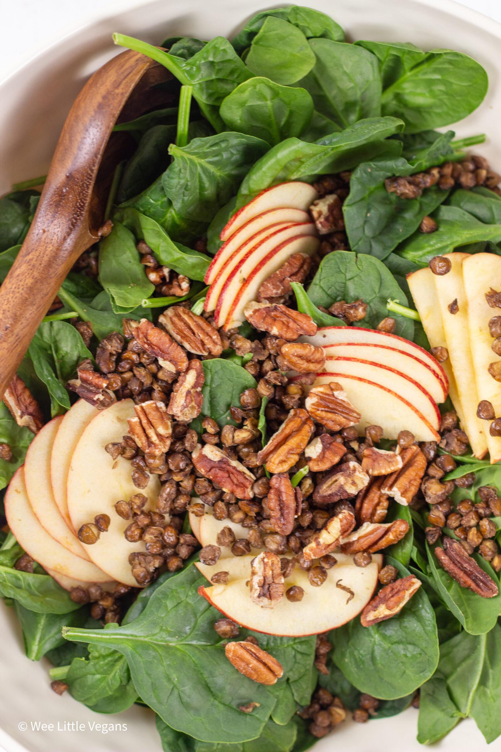 Close up of Apple Pecan Greens Salad in a large bowl with a wooden spoon to the side.