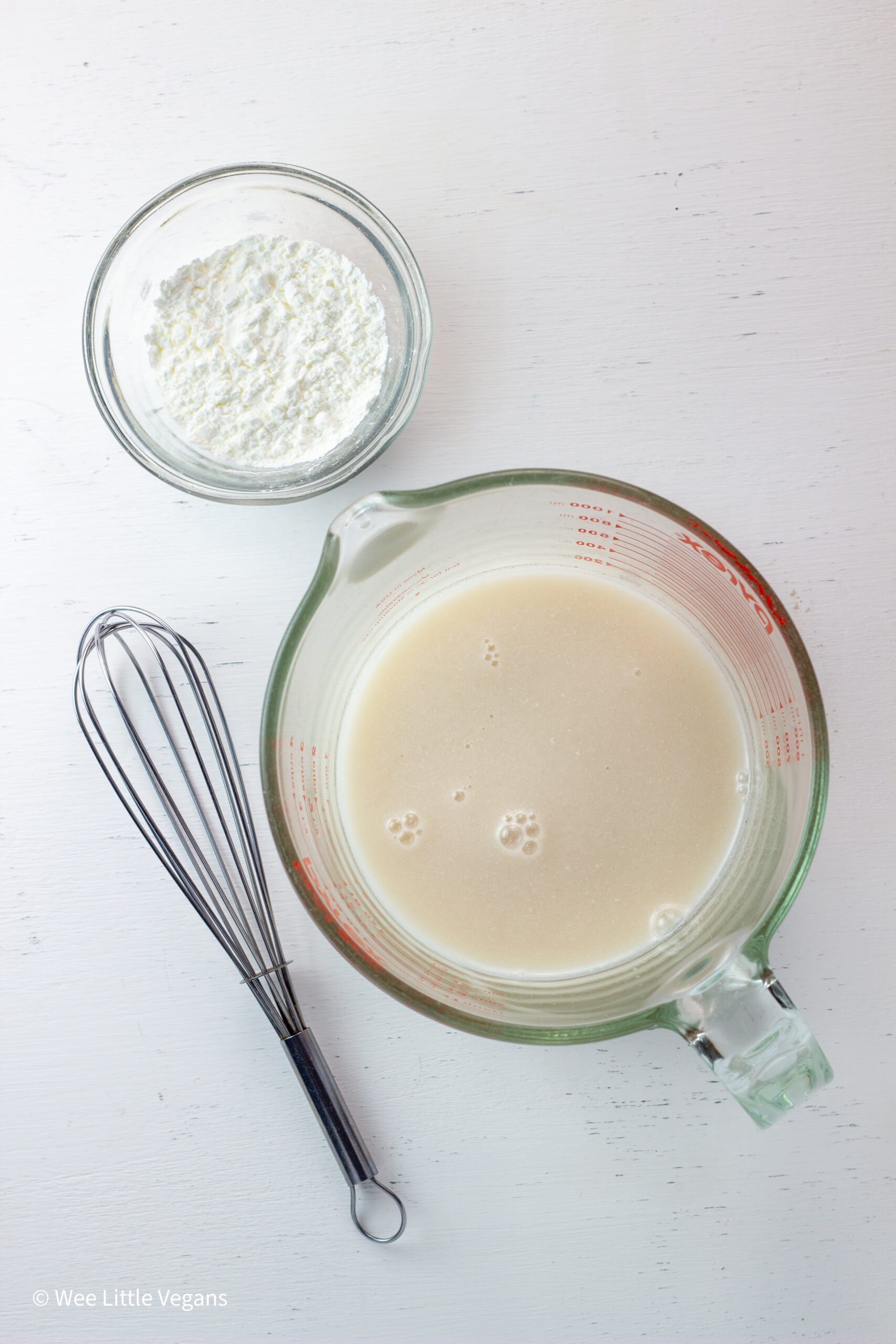 Overhead shot of a measuring cup of banana milk, a small bowl of cornstarch and a whisk.
