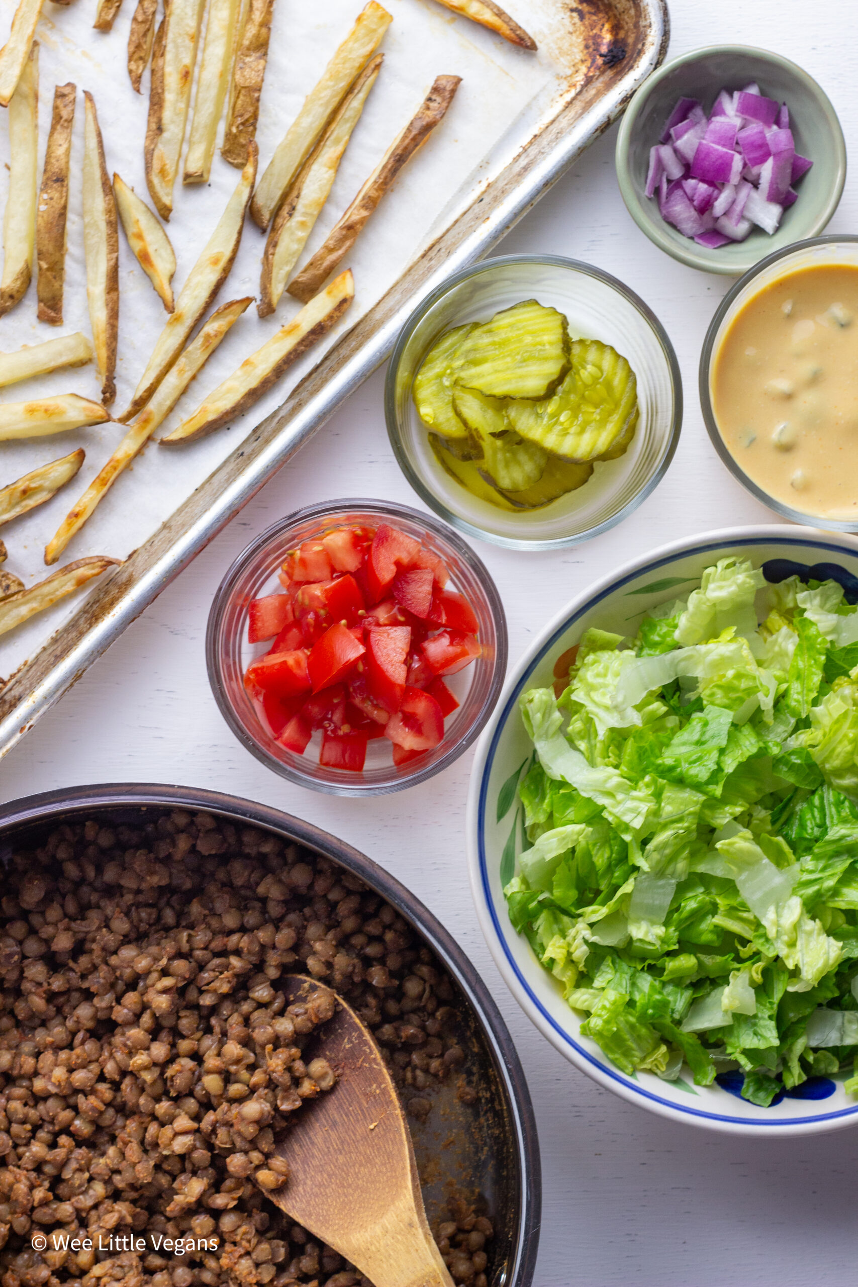 Overhead shot of lentils in a pan, small bowls of lettuce, tomatoes, pickles, special sauce, diced red onion and a sheet pan of fries.