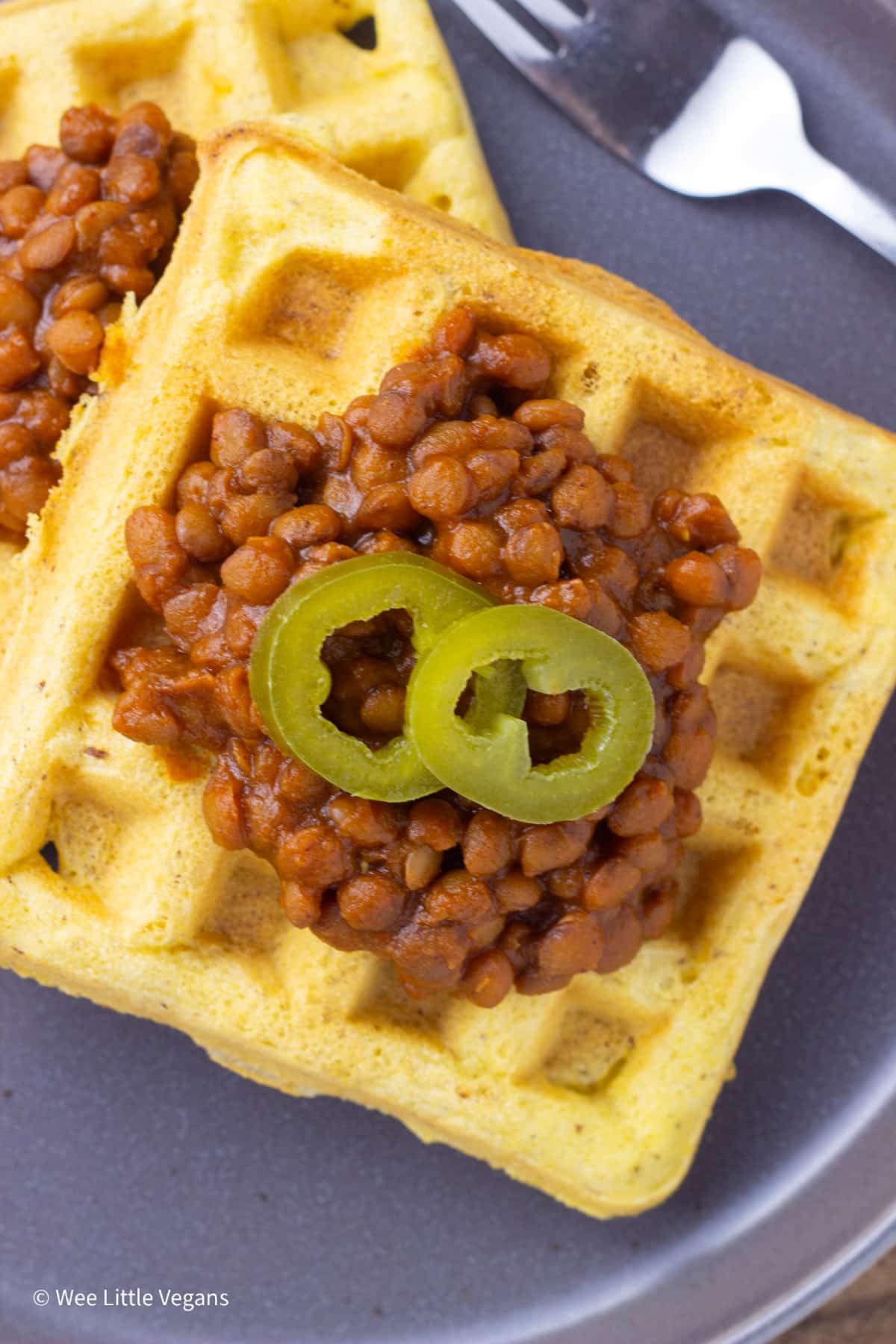 Overhead close up view of a cornbread waffle topped with barbecue lentils and two pickled jalapeño slices.