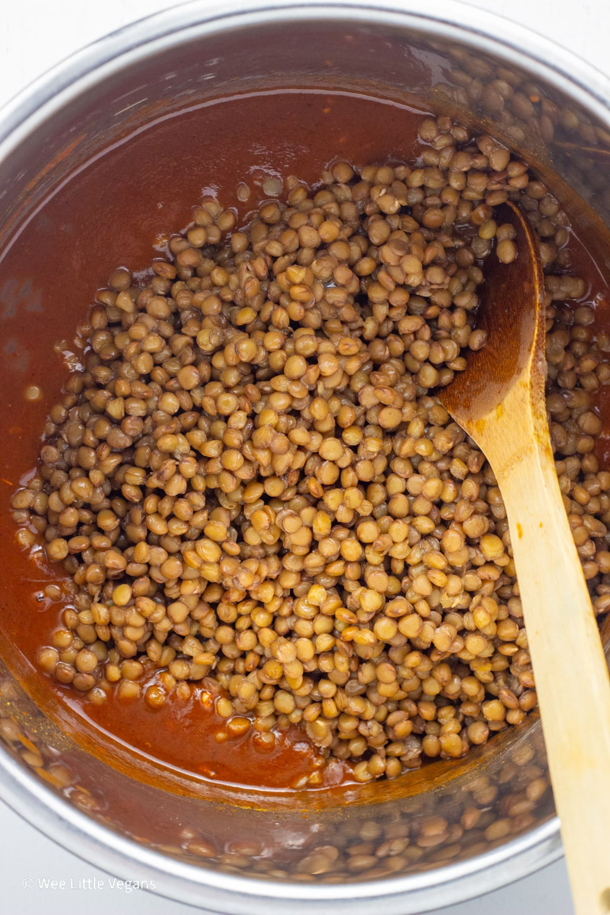 Overhead view of a pot with red sauce and cooked lentils before mixing together. There is a wooden spoon resting in the pot.