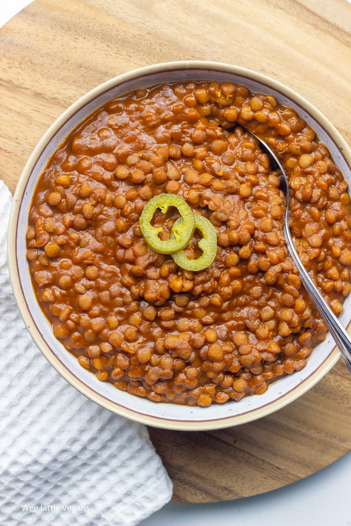 Overhead shot of a bowl of barbecue lentils with slices of pickled jalapeño on top. There is a spoon in the bowl and a white towel to the side of the dish.