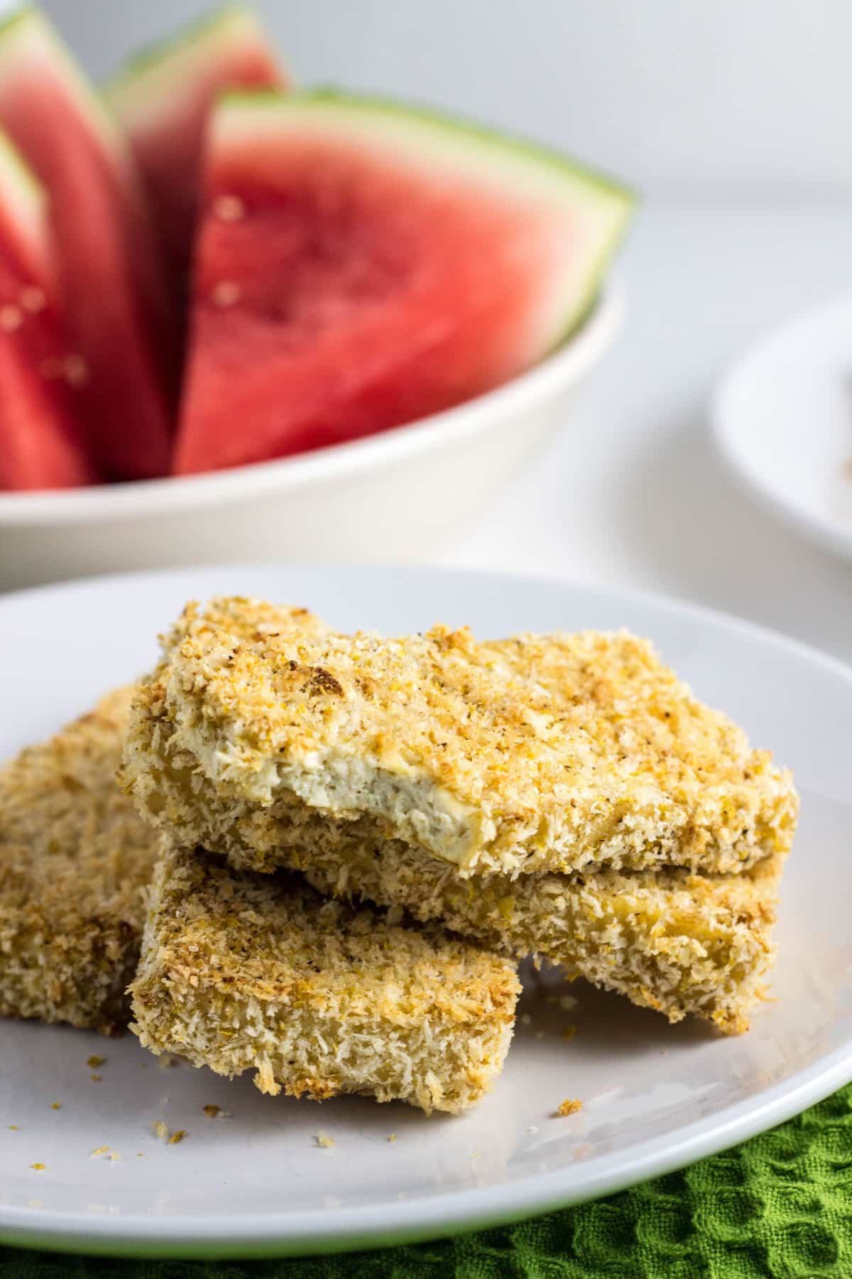 Close up of Breaded Tofu Sticks on a small plate. There is a bite revealing the interior texture. There is a bowl of watermelon slices in the background.